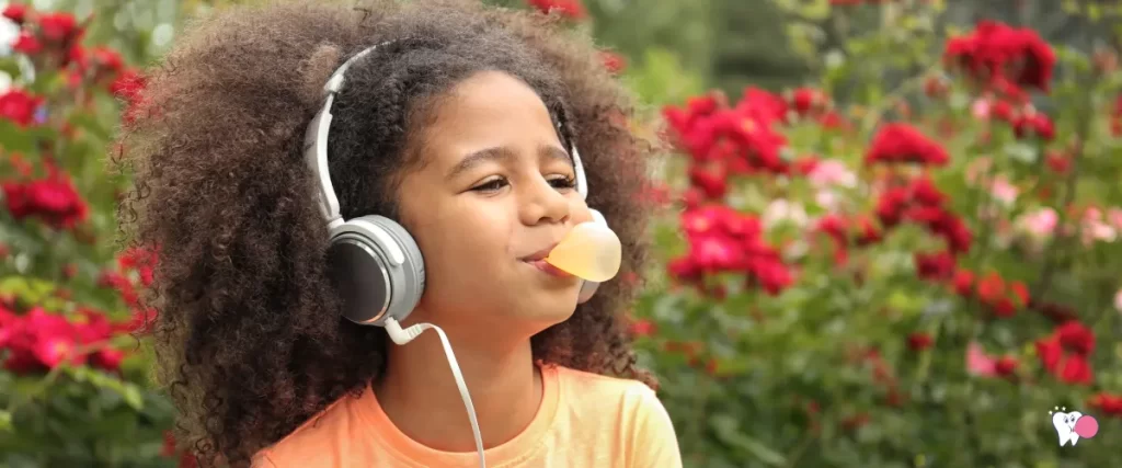 An Afro-american girl child is pleasantly blowing a chewing gum while her headphones on in a garden | Source: shutterstock | For the article: What happens if you accidently swallow gum? | For the website: healthchewinggum.com (Health Chewing Gum)