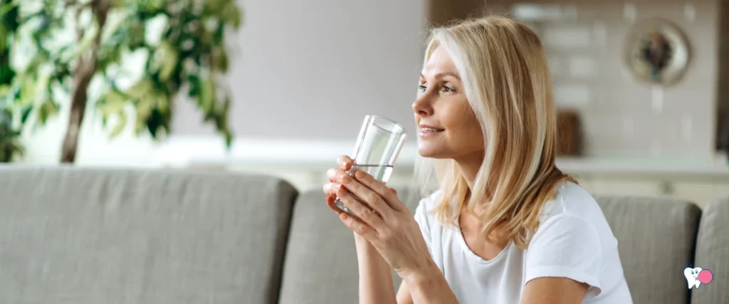 An elder woman is sitting in her living room on a grey sofa and drinking water | For the article: What Causes Dry Mouth and How to Relieve it? | For the website: healthchewinggum.com (Health Chewing Gum)