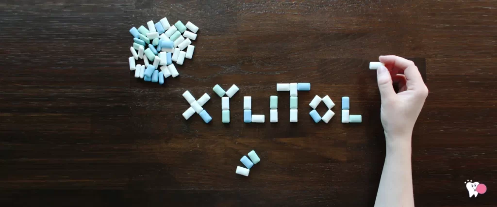Against a black background there are xylitol chewing gums and a hand is trying to use the chewing gums to write the word Xylitol with them | For the article: Why do you need xylitol in your chewing gum | For the website: healthchewinggum.com (Health Chewing Gum)