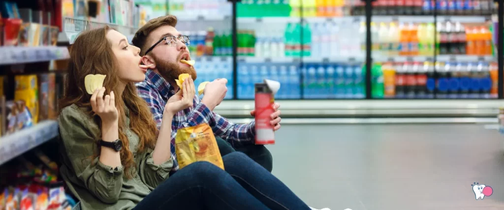 A young male and a young female can be seen sitting along the grossery line in a shop whille they pig out on the snacks | For the article: Is Chewing Gum Better Than Snacking? | For the website: healthchewinggum.com (Health Chewing Gum)