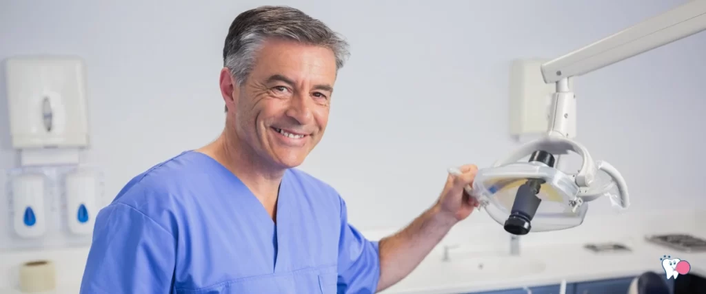 The image shows a middle aged male dentist smiling at the camera while he is stanidng in the clinic | For the article: When do you need to see your dentist? | For the website: healthchewinggum.com (Health Chewing Gum)